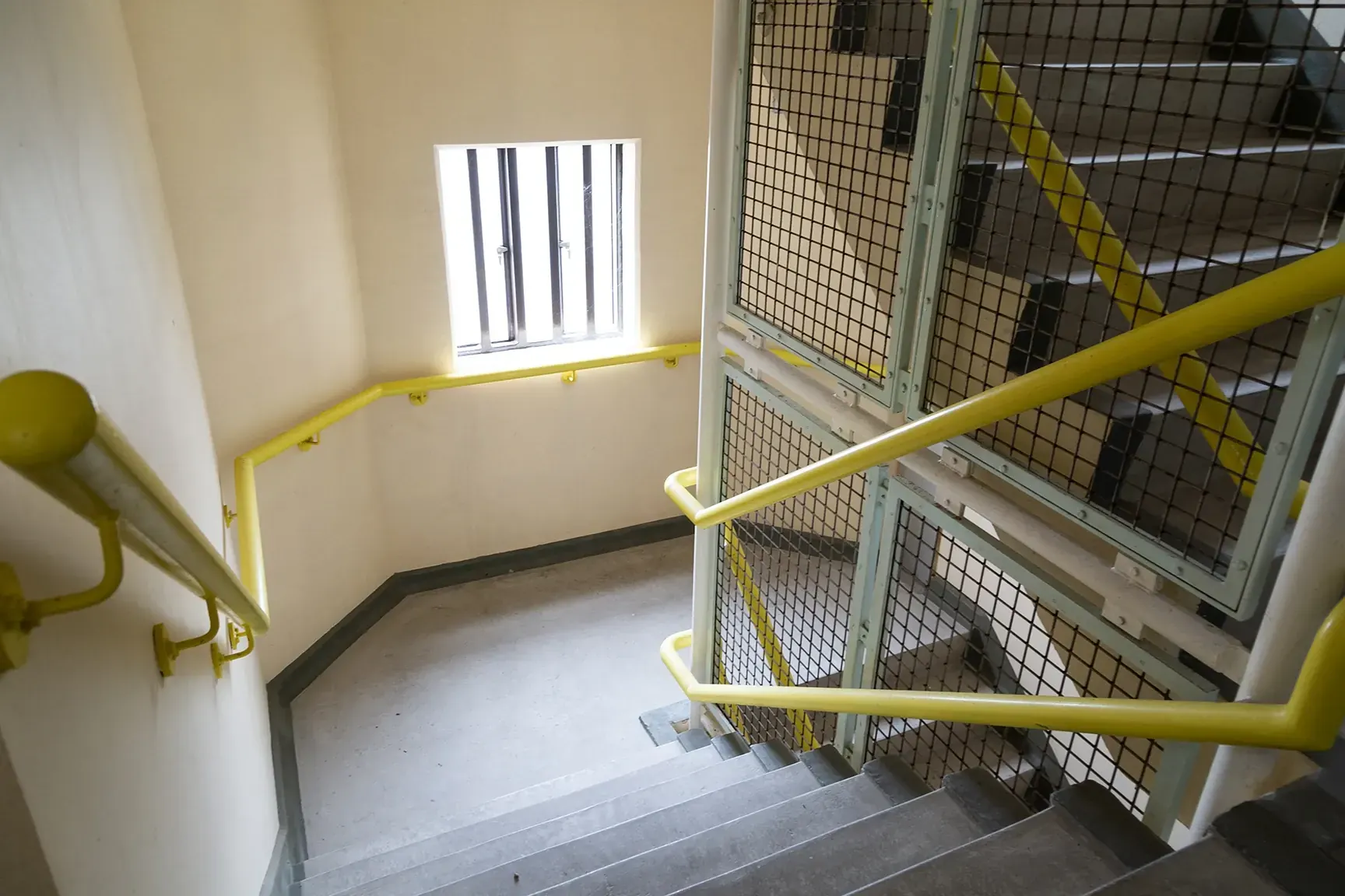 Prison stairwell with yellow handrails, wire-mesh cage and barred landing window