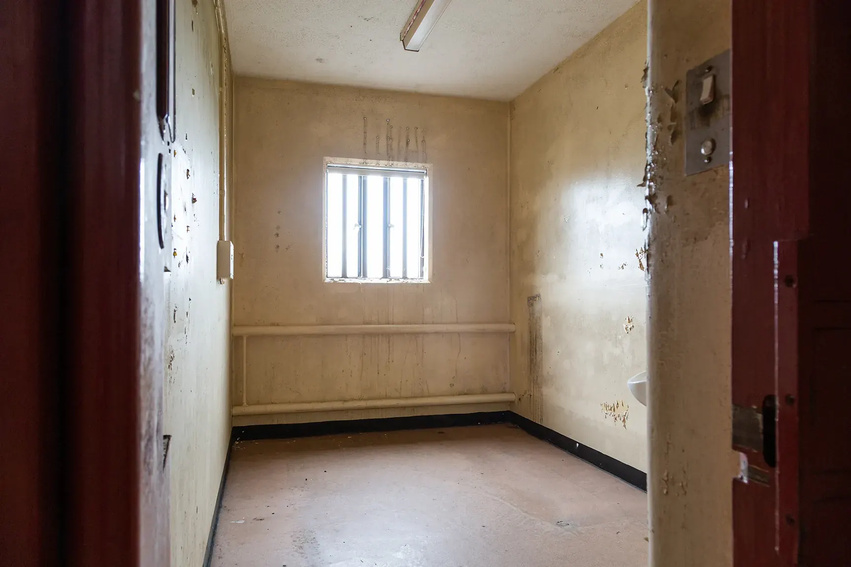Empty prison cell with barred window, exposed pipework and worn painted walls