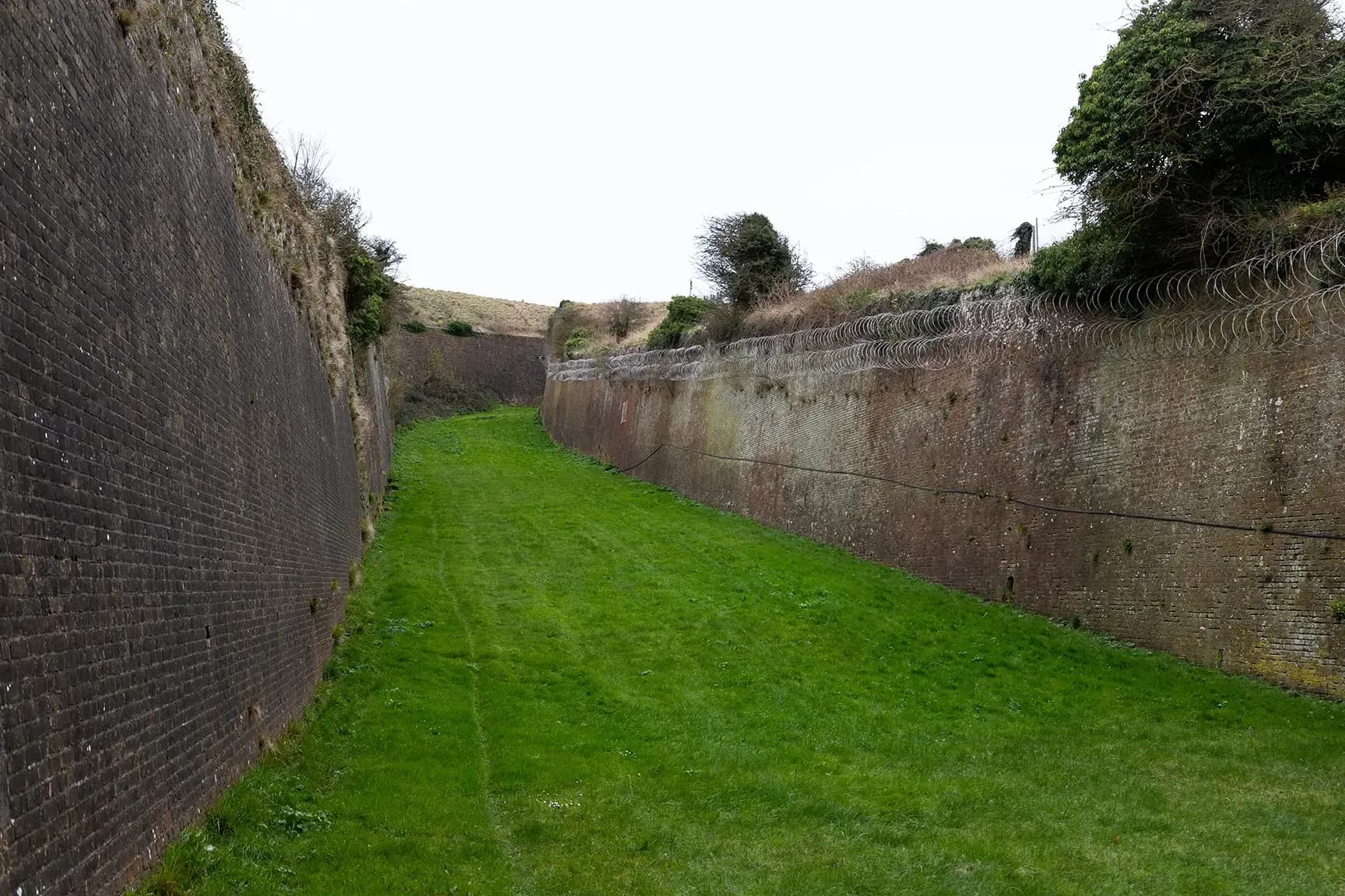 Grassy path between high brick walls topped with concertina wire